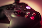 A close-up of a video game controller with a focus on the buttons and joystick. The buttons are marked with letters and are lit up in different colors against a dark surface. The background is blurred, creating a moody atmosphere.