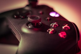 A close-up of a video game controller with a focus on the buttons and joystick. The buttons are marked with letters and are lit up in different colors against a dark surface. The background is blurred, creating a moody atmosphere.