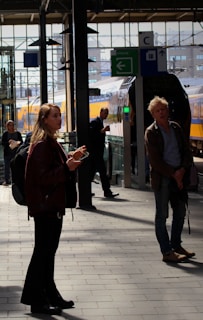 Several people are standing on a train platform, some appearing to wait while others are engaged with their phones. A train is visible in the background, and various signs are present overhead. The station has a modern, industrial look with metal beams and glass.