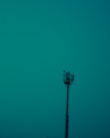A tall communication tower stands prominently against a teal sky, with visible antennas and equipment at the top.