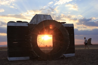 A large, artistic wooden camera structure stands on a barren landscape, framing a vibrant sunset. Sun rays illuminate the clouds in the sky, casting a warm glow over the scene. A person is walking on the right side of the image, adding a sense of scale to the camera sculpture and the landscape.