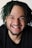 A person with dreadlocks smiles broadly, showcasing a joyful expression. The hairstyle includes green-dyed locks, and the individual has facial hair including a beard and mustache.