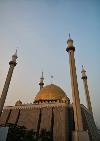 A large mosque with a grand golden dome and four tall minarets reaching towards a clear sky. The architectural style is ornate, with intricate details and geometric patterns visible on the facade. The setting sun casts a warm, golden hue over the structure.