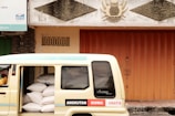 A cream-colored van partially loaded with large white bags is parked beside a building with an orange metal door. The building features decorative reliefs and a bank sign is visible at the top left. The words 'ANGKUTAN SISWA GRATIS' are printed on the side of the van.