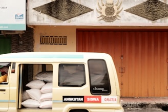A cream-colored van partially loaded with large white bags is parked beside a building with an orange metal door. The building features decorative reliefs and a bank sign is visible at the top left. The words 'ANGKUTAN SISWA GRATIS' are printed on the side of the van.