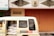 A cream-colored van partially loaded with large white bags is parked beside a building with an orange metal door. The building features decorative reliefs and a bank sign is visible at the top left. The words 'ANGKUTAN SISWA GRATIS' are printed on the side of the van.