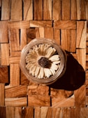 A dried flower with beige petals and a brown center is placed inside a clear glass container. The container is positioned on a wooden surface with square tiles of varying shades of brown, creating a warm and rustic atmosphere.