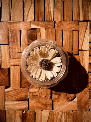 A dried flower with beige petals and a brown center is placed inside a clear glass container. The container is positioned on a wooden surface with square tiles of varying shades of brown, creating a warm and rustic atmosphere.