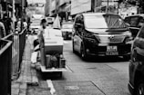 A bustling urban street with a person on a cart carrying packages, navigating through heavy traffic. A large black van is prominently visible, along with other vehicles and pedestrians on the sidewalk. The scene is set in a city environment with tall buildings and store signs.