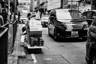 A bustling urban street with a person on a cart carrying packages, navigating through heavy traffic. A large black van is prominently visible, along with other vehicles and pedestrians on the sidewalk. The scene is set in a city environment with tall buildings and store signs.