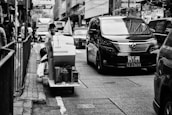 A bustling urban street with a person on a cart carrying packages, navigating through heavy traffic. A large black van is prominently visible, along with other vehicles and pedestrians on the sidewalk. The scene is set in a city environment with tall buildings and store signs.