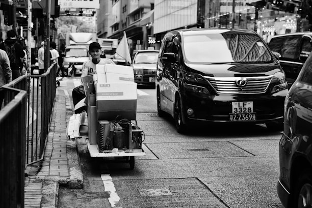 A bustling urban street with a person on a cart carrying packages, navigating through heavy traffic. A large black van is prominently visible, along with other vehicles and pedestrians on the sidewalk. The scene is set in a city environment with tall buildings and store signs.