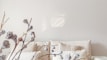 A minimalist interior featuring a beige couch adorned with several matching pillows. In the foreground, there are delicate dried branches with small blossoms, adding a natural element to the scene. The setting conveys a calm and serene atmosphere with soft lighting.