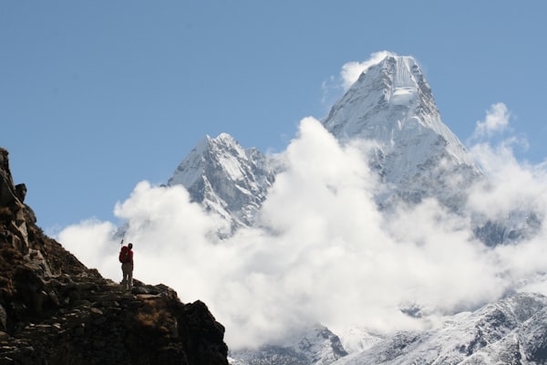 A person standing on a rocky mountain, observing snowy peaks through thick clouds.