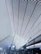 A modern interior with a high, angular ceiling featuring vertical slats creating a striped pattern. The space is illuminated with cool white lighting, enhancing the sleek, futuristic design. On one of the walls, signage displays the words 'Tea Geek Lab' and 'Shenzhen' alongside a circular logo. The environment has a metallic and minimalist aesthetic.