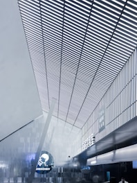 A modern interior with a high, angular ceiling featuring vertical slats creating a striped pattern. The space is illuminated with cool white lighting, enhancing the sleek, futuristic design. On one of the walls, signage displays the words 'Tea Geek Lab' and 'Shenzhen' alongside a circular logo. The environment has a metallic and minimalist aesthetic.