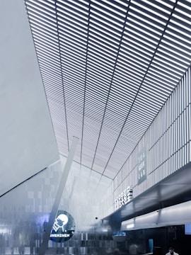 A modern interior with a high, angular ceiling featuring vertical slats creating a striped pattern. The space is illuminated with cool white lighting, enhancing the sleek, futuristic design. On one of the walls, signage displays the words 'Tea Geek Lab' and 'Shenzhen' alongside a circular logo. The environment has a metallic and minimalist aesthetic.