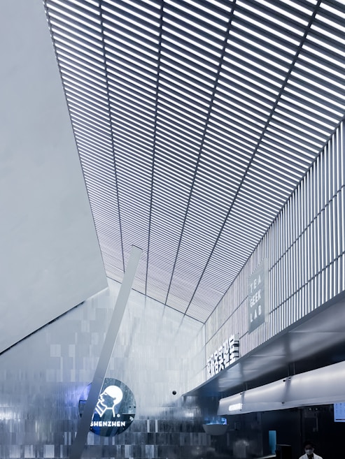 A modern interior with a high, angular ceiling featuring vertical slats creating a striped pattern. The space is illuminated with cool white lighting, enhancing the sleek, futuristic design. On one of the walls, signage displays the words 'Tea Geek Lab' and 'Shenzhen' alongside a circular logo. The environment has a metallic and minimalist aesthetic.