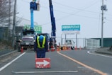 A construction worker dressed in blue uniform and yellow safety vest is bending over a red sign with a white arrow. Behind him, there are utility trucks with cranes raised and another worker in a blue uniform working on an electrical pole. The road is partially blocked with orange traffic cones, and there is a sign in the background, written in a foreign language, advertising a clinic.