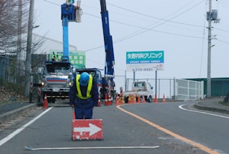 A construction worker dressed in blue uniform and yellow safety vest is bending over a red sign with a white arrow. Behind him, there are utility trucks with cranes raised and another worker in a blue uniform working on an electrical pole. The road is partially blocked with orange traffic cones, and there is a sign in the background, written in a foreign language, advertising a clinic.