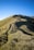 A trekking path leads up a grassy hill with several hikers visible along the trail. The sky is clear and blue, creating a bright and serene atmosphere. The terrain is gently sloping with patches of shadows cast by the uneven ground and surrounding vegetation.