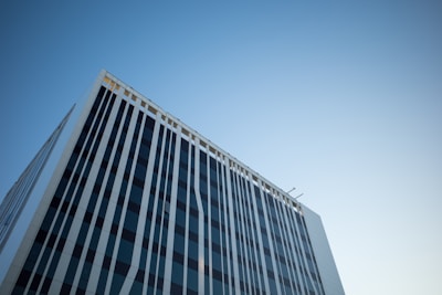A tall modern building with a sleek design stands against a clear blue sky. The facade features vertical lines with a pattern of alternating dark and light shades. The architectural style is contemporary, with clean lines and minimalistic detail.
