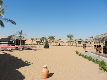 A desert setting with a clear blue sky, featuring several palm trees and thatched-roof huts. There are seating arrangements on the sand, including low tables and cushions. A lone person is walking near the wooden structures, and a decorated tree is prominently placed amid the sandy expanse. Terra cotta pots and green shrubs add detail to the scene.