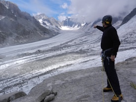 A person wearing climbing gear is standing on a rocky surface, pointing towards a vast glacier surrounded by steep mountains under a partly cloudy sky.