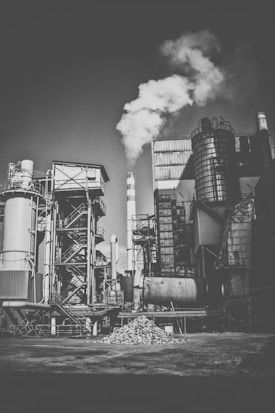 A monochrome photograph depicting an industrial facility with various metal structures and pipes. Smoke is billowing from a tall chimney, indicating active industrial processes. The ground is piled with rubble or gravel, and the overall setting suggests a factory or plant environment.