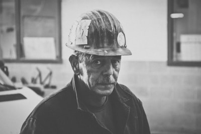 A black and white photo of a man wearing a safety helmet and a work jacket. The background shows an industrial setting with blurred machinery and office windows.