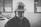 A black and white photo of a man wearing a safety helmet and a work jacket. The background shows an industrial setting with blurred machinery and office windows.