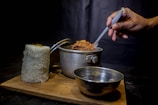 A hand holding a spoon is stirring a pot of food, which is placed on a wooden board. Next to the pot, there is a tall cylindrical mound of white rice and an empty metal bowl. The background is dark, creating a contrast with the bright colors of the food.
