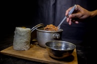 A hand holding a spoon is stirring a pot of food, which is placed on a wooden board. Next to the pot, there is a tall cylindrical mound of white rice and an empty metal bowl. The background is dark, creating a contrast with the bright colors of the food.