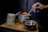 A hand holding a spoon is stirring a pot of food, which is placed on a wooden board. Next to the pot, there is a tall cylindrical mound of white rice and an empty metal bowl. The background is dark, creating a contrast with the bright colors of the food.