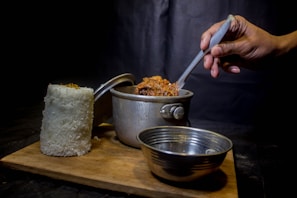 A hand holding a spoon is stirring a pot of food, which is placed on a wooden board. Next to the pot, there is a tall cylindrical mound of white rice and an empty metal bowl. The background is dark, creating a contrast with the bright colors of the food.