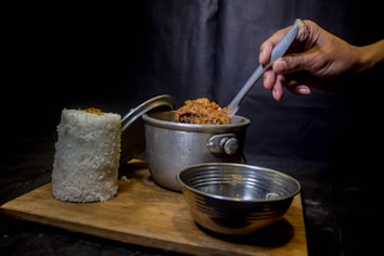 A hand holding a spoon is stirring a pot of food, which is placed on a wooden board. Next to the pot, there is a tall cylindrical mound of white rice and an empty metal bowl. The background is dark, creating a contrast with the bright colors of the food.