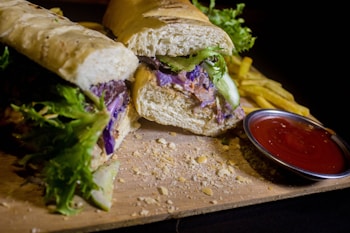 A sandwich stuffed with various fresh ingredients such as lettuce and purple cabbage, placed on a wooden board. Next to the sandwich are crispy golden fries and a small bowl of red ketchup, likely for dipping. The surrounding crumbs and casual placement suggest a casual dining setting.