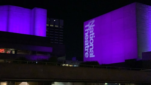 A large building is illuminated with purple light at night. The words 'National Theatre' are prominently projected onto one side of the structure.