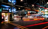 An urban street scene at night featuring illuminated storefronts, including a shop named 'Coffee Criminals.' Bright, colorful light trails suggestive of moving vehicles create dynamic lines across the scene. The area is busy with traffic, and the ambiance is enhanced by overhead streetlights and glowing signs.