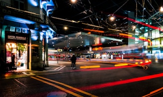 An urban street scene at night featuring illuminated storefronts, including a shop named 'Coffee Criminals.' Bright, colorful light trails suggestive of moving vehicles create dynamic lines across the scene. The area is busy with traffic, and the ambiance is enhanced by overhead streetlights and glowing signs.