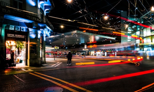 An urban street scene at night featuring illuminated storefronts, including a shop named 'Coffee Criminals.' Bright, colorful light trails suggestive of moving vehicles create dynamic lines across the scene. The area is busy with traffic, and the ambiance is enhanced by overhead streetlights and glowing signs.