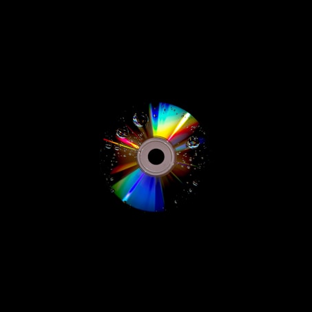 A compact disc with a colorful spectrum of light reflecting on its surface. The disc is adorned with water droplets, enhancing the rainbow effect, and is set against a dark, black background.