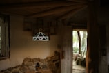 A cozy interior with rustic decor, featuring a wooden ceiling and walls partially made of stone. A geometric, decorative pendant light hangs from the ceiling, illuminated softly. The room includes an open wooden door leading to an outdoor area with trees and wooden seating. A small lantern sits on a table beneath the light fixture.