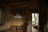 A cozy interior with rustic decor, featuring a wooden ceiling and walls partially made of stone. A geometric, decorative pendant light hangs from the ceiling, illuminated softly. The room includes an open wooden door leading to an outdoor area with trees and wooden seating. A small lantern sits on a table beneath the light fixture.