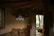 A cozy interior with rustic decor, featuring a wooden ceiling and walls partially made of stone. A geometric, decorative pendant light hangs from the ceiling, illuminated softly. The room includes an open wooden door leading to an outdoor area with trees and wooden seating. A small lantern sits on a table beneath the light fixture.