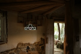 A cozy interior with rustic decor, featuring a wooden ceiling and walls partially made of stone. A geometric, decorative pendant light hangs from the ceiling, illuminated softly. The room includes an open wooden door leading to an outdoor area with trees and wooden seating. A small lantern sits on a table beneath the light fixture.