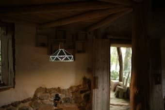 A cozy interior with rustic decor, featuring a wooden ceiling and walls partially made of stone. A geometric, decorative pendant light hangs from the ceiling, illuminated softly. The room includes an open wooden door leading to an outdoor area with trees and wooden seating. A small lantern sits on a table beneath the light fixture.