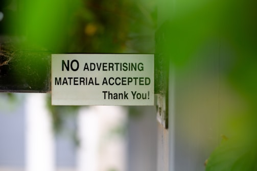A sign attached to a door or wall states 'NO ADVERTISING MATERIAL ACCEPTED Thank You!' with a background partially obscured by green leaves, creating a soft focus effect.