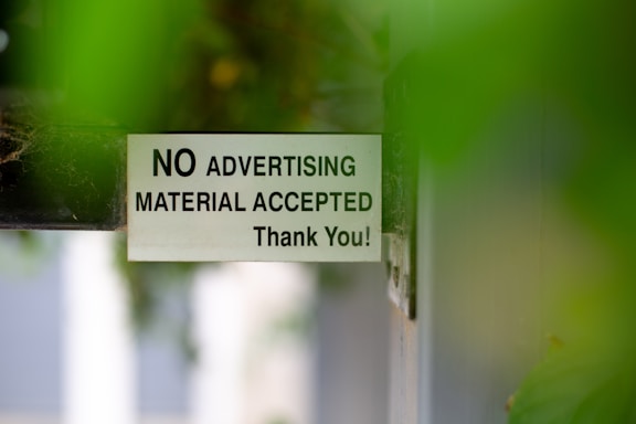 A sign attached to a door or wall states 'NO ADVERTISING MATERIAL ACCEPTED Thank You!' with a background partially obscured by green leaves, creating a soft focus effect.