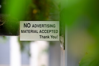 A sign attached to a door or wall states 'NO ADVERTISING MATERIAL ACCEPTED Thank You!' with a background partially obscured by green leaves, creating a soft focus effect.